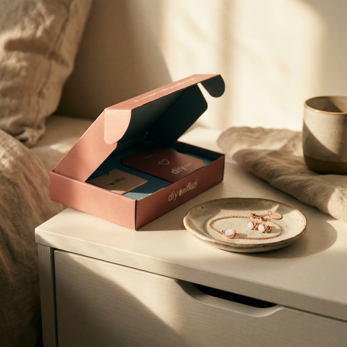 Warm morning sunlight on a bedside table with a DIY by MILKIES kit box beside rose-gold nursing jewelry, including a bracelet and earrings with milky white stones.