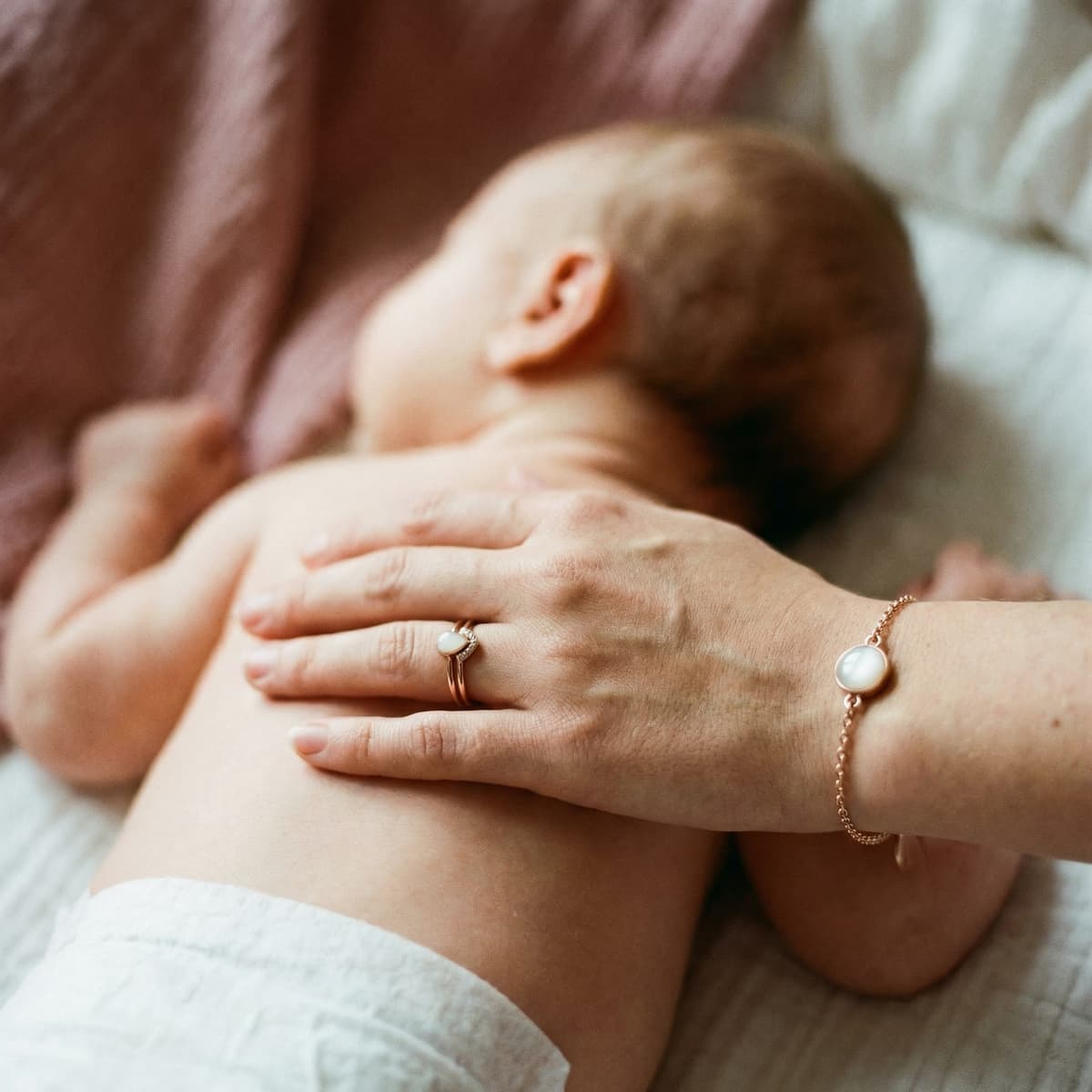Mother gently rests her hand on a sleeping newborn, wearing a rose-gold nursing keepsake ring and matching bracelet with a milky-white stone.