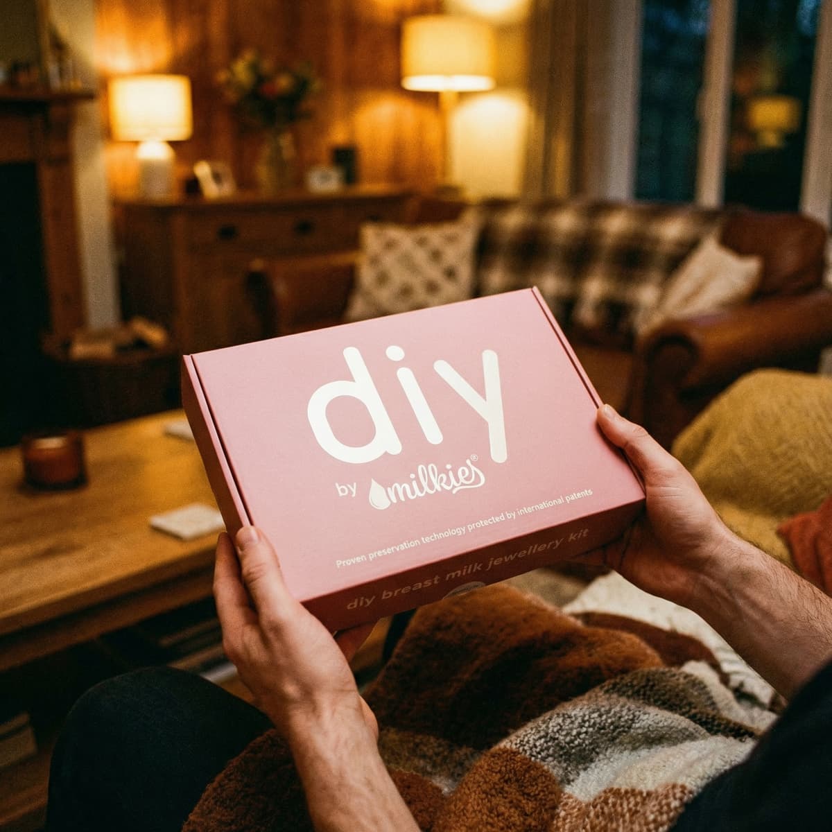 Hands holding a pink DIY by MILKIES breast milk jewellery kit box in a cozy living room, a thoughtful push present for wife to create a keepsake necklace, ring or earrings at home.