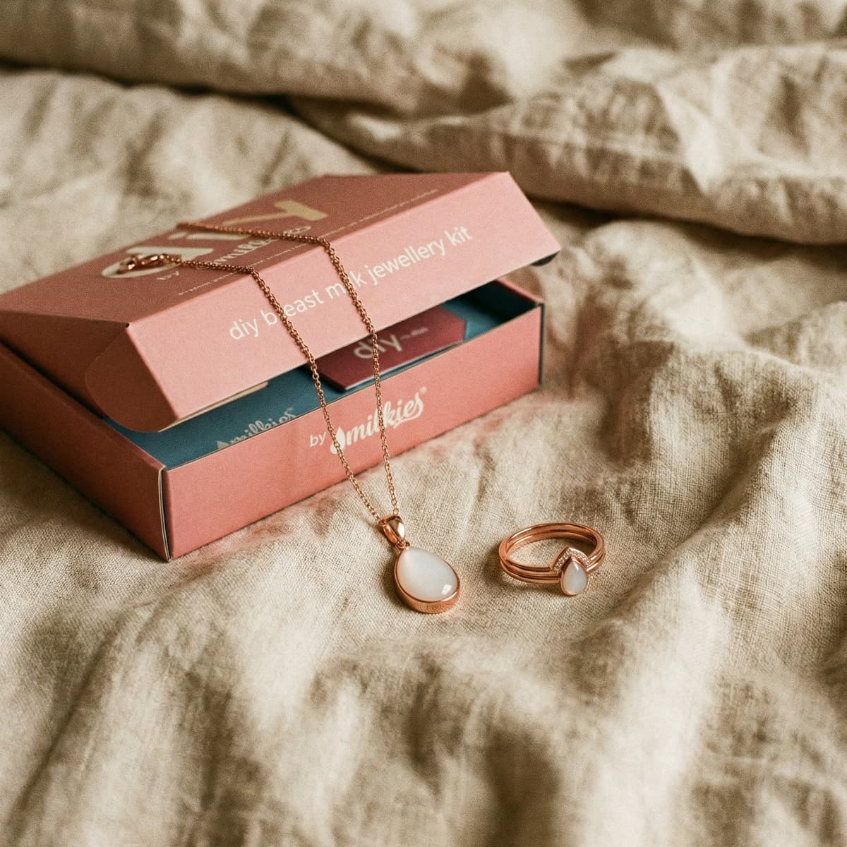 Elegant DIY by MILKIES breast milk jewelry kit box on a soft bed, a thoughtful push present for wife, featuring a rose-gold teardrop pendant necklace and matching ring keepsake.