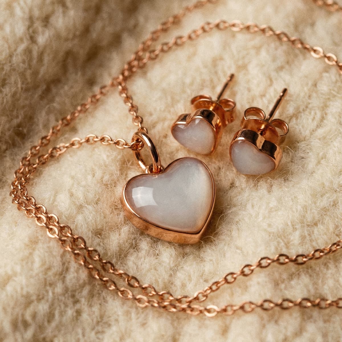 Rose gold heart pendant necklace and matching heart stud earrings with milky white stones on soft beige fabric, a sentimental keepsake jewelry set ideal as a weaning gift.