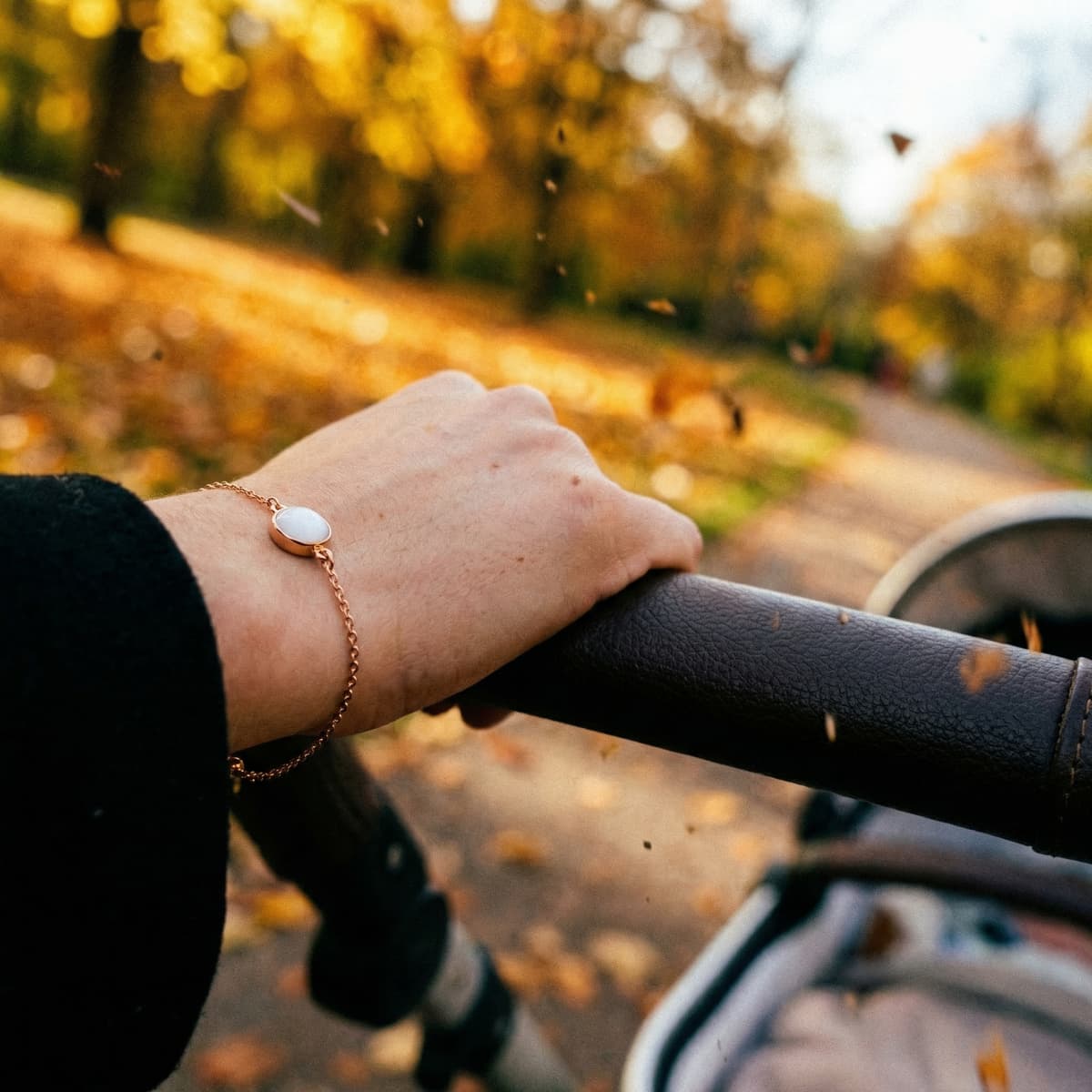 Rose-gold bracelet with a white resin stone worn on a hand pushing a stroller along an autumn path, representing a 1 year breastfeeding keepsake jewelry memory.