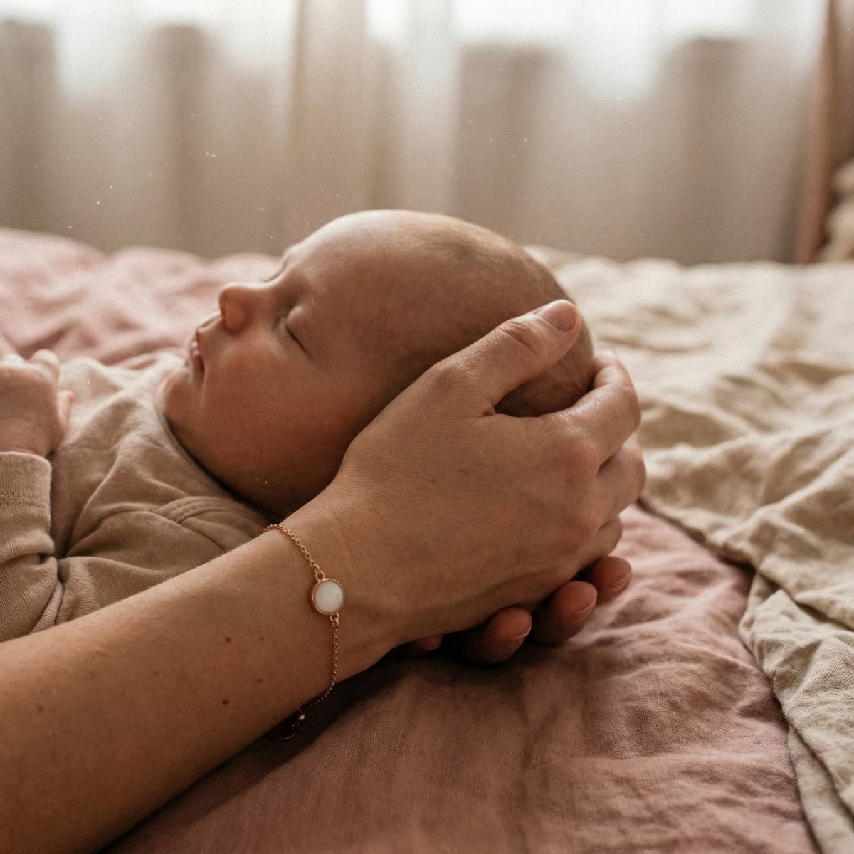 Mother cradling a sleeping newborn on a bed while wearing a delicate keepsake bracelet, illustrating why a breastmilk bracelet diy making kit is a meaningful at-home way to create a personal memory.