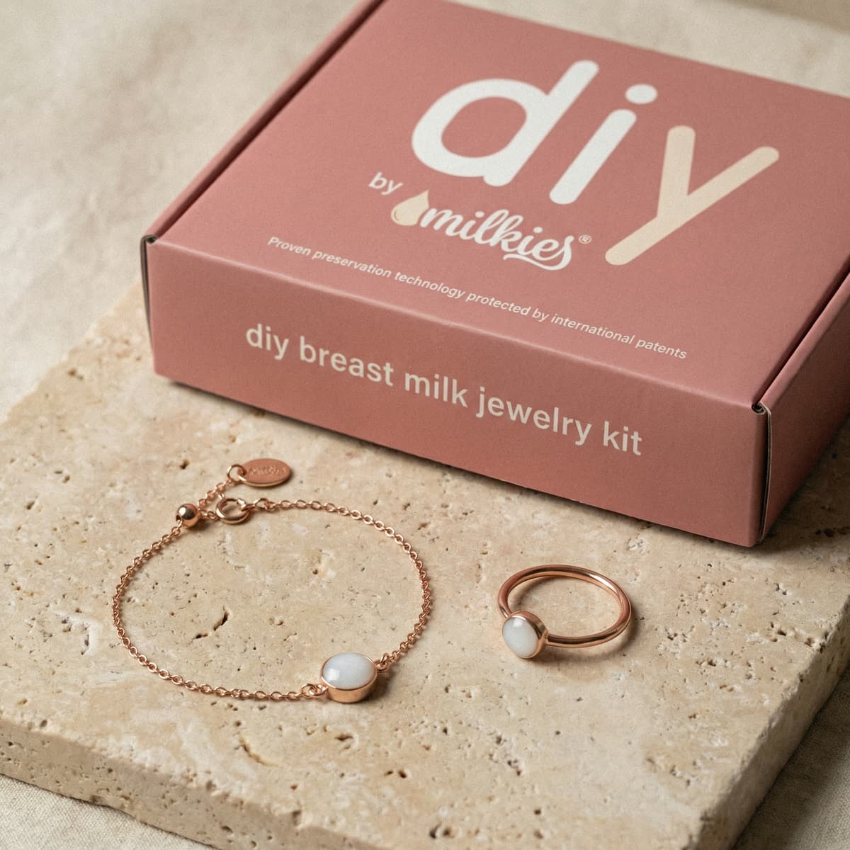 Rose-gold jewelry displayed on a stone surface beside a DIY by MILKIES at-home breastmilk bracelet making kit for creating breast milk jewelry keepsakes.