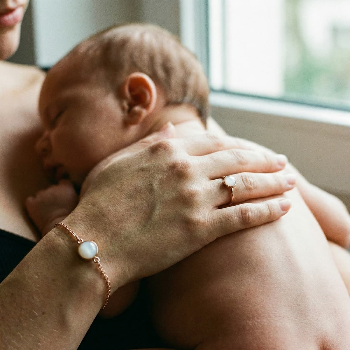 Mother cuddling a sleeping newborn by a window, wearing matching keepsake jewelry, illustrating why a breastmilk bracelets diy making kit is a meaningful at-home way to preserve precious moments.