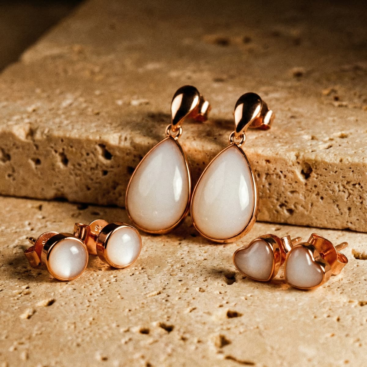 Rose gold breastmilk keepsake earrings on a beige stone surface, featuring teardrop dangle and round and heart stud styles made with a breastmilk earring diy making kit