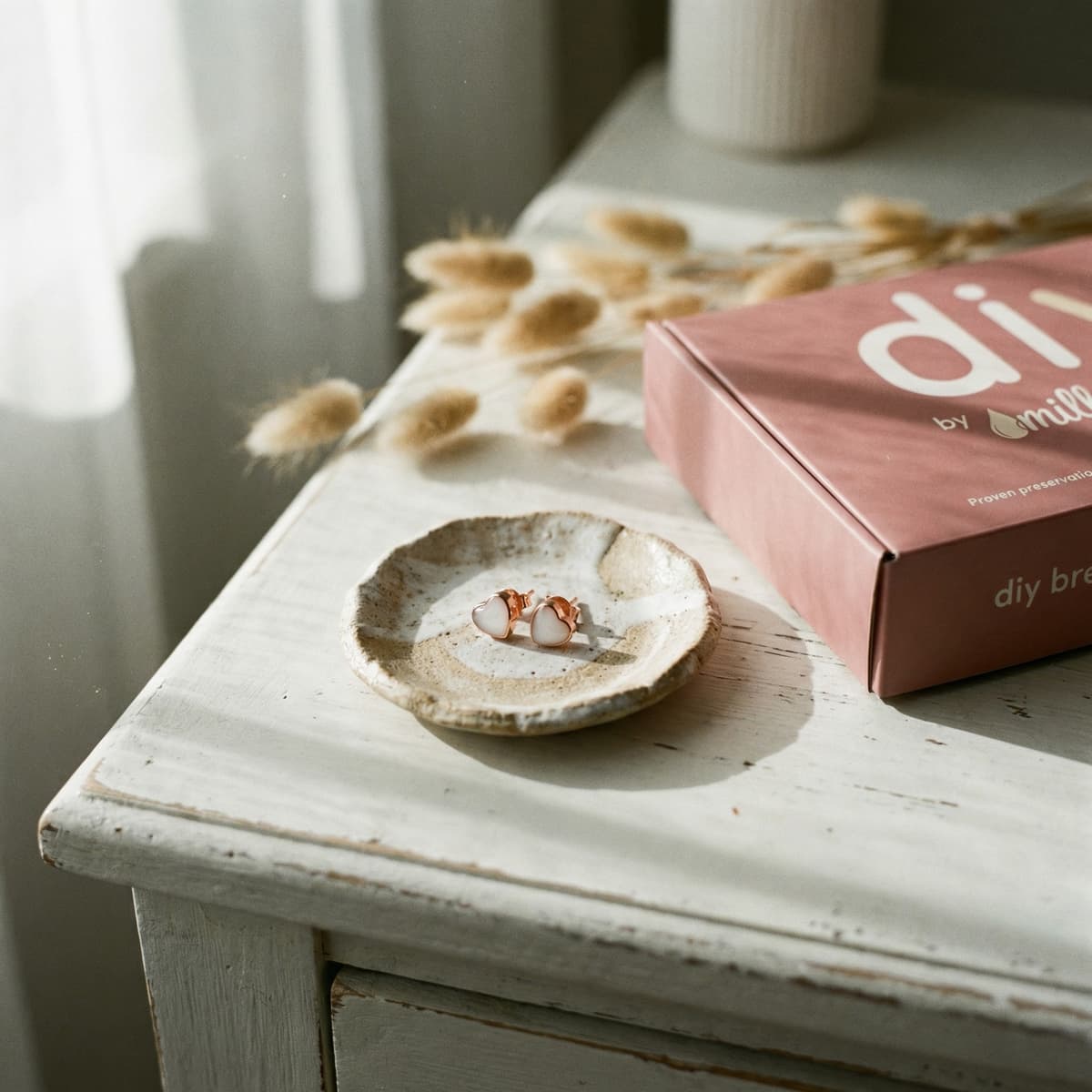 Rose-gold stud earrings with milky white stones on a rustic ceramic dish beside a pink DIY by MILKIES box, featuring a breastmilk earring making kit in soft natural light.
