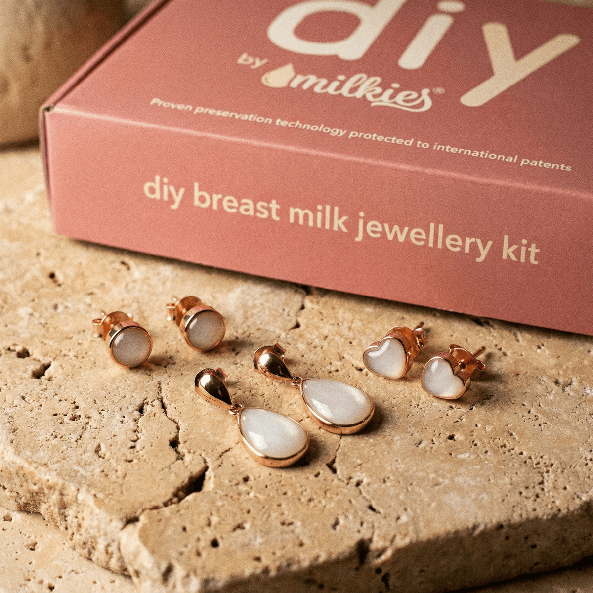 Rose-gold DIY by MILKIES breastmilk jewellery kit box displayed with white resin teardrop and stud earrings, showing a breastmilk earrings diy making kit on a stone surface.