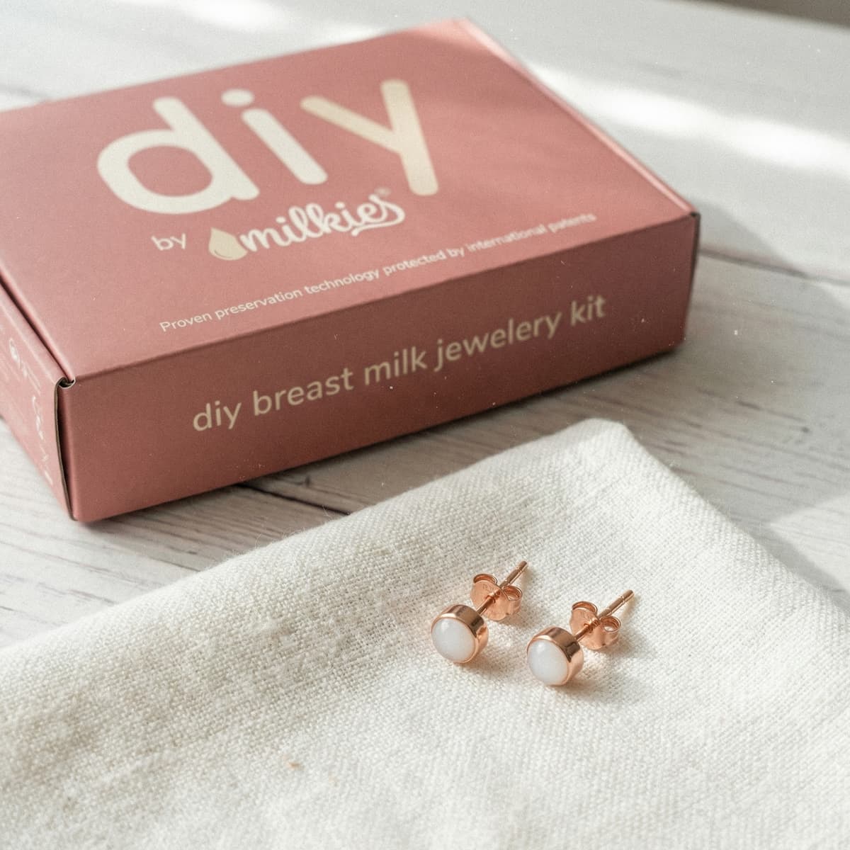 Rose-gold stud earrings on a linen cloth next to the DIY by MILKIES box, showing an at-home breastmilk earrings making kit for creating breast milk jewelry keepsakes.