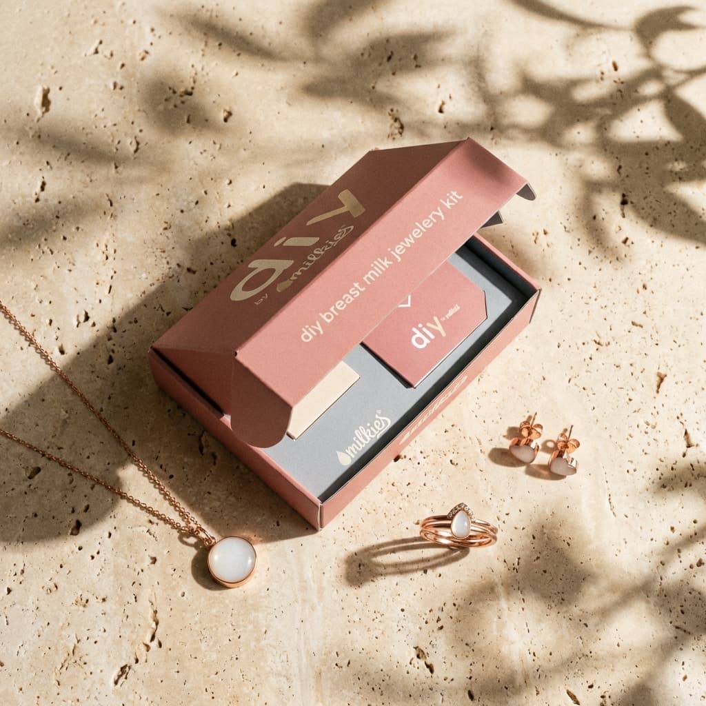 Open pink DIY by MILKIES breastmilk jewellery making kit displayed on a stone surface with a rose-gold necklace, ring, and matching earrings featuring milky white resin keepsakes.