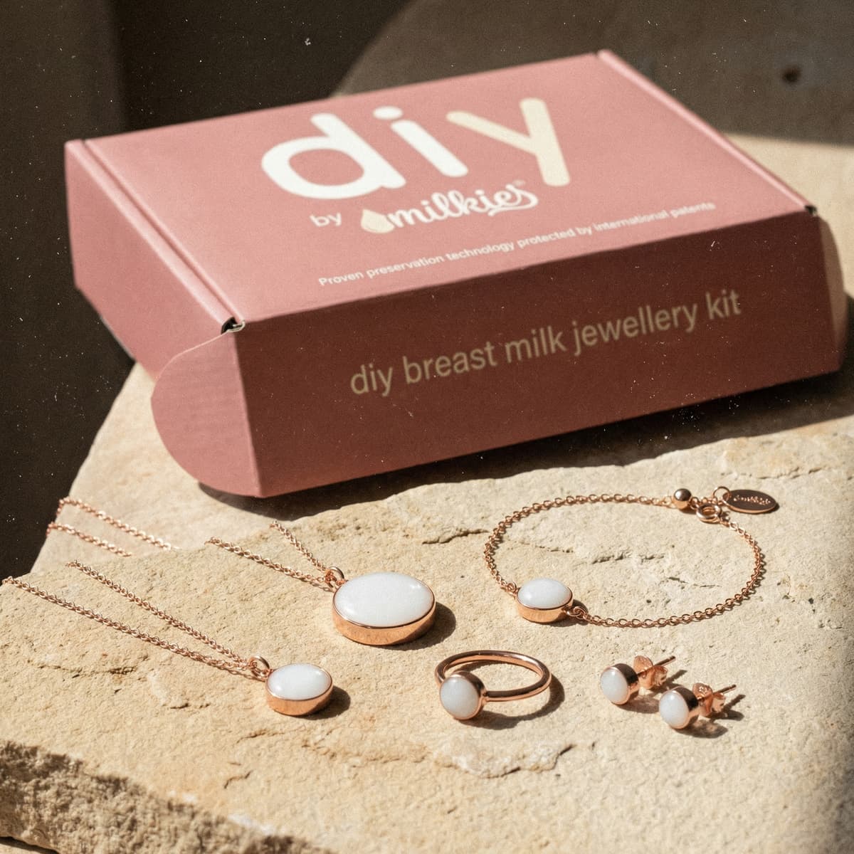 Rose-gold DIY by MILKIES box with finished keepsakes from a breastmilk jewelry diy making kit, showing white resin pendant necklaces, a ring, bracelet, and stud earrings displayed on stone.