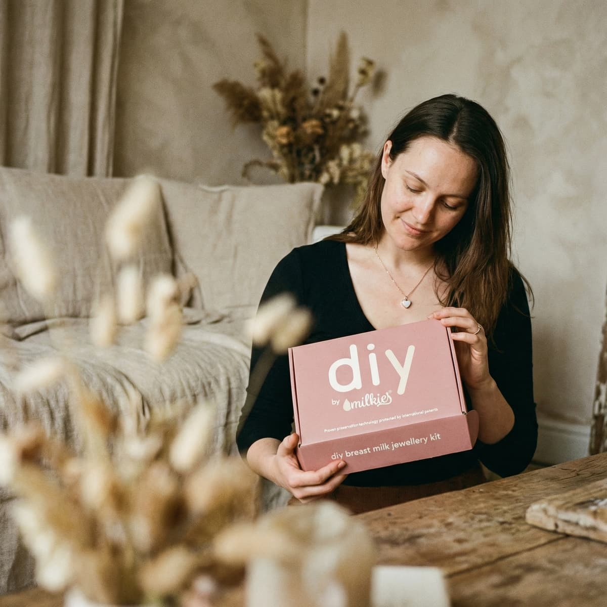 Mother at home holding a DIY by MILKIES box with a breastmilk jewelry making kit, highlighting an easy at-home way to create a meaningful keepsake with patented preservation technology.