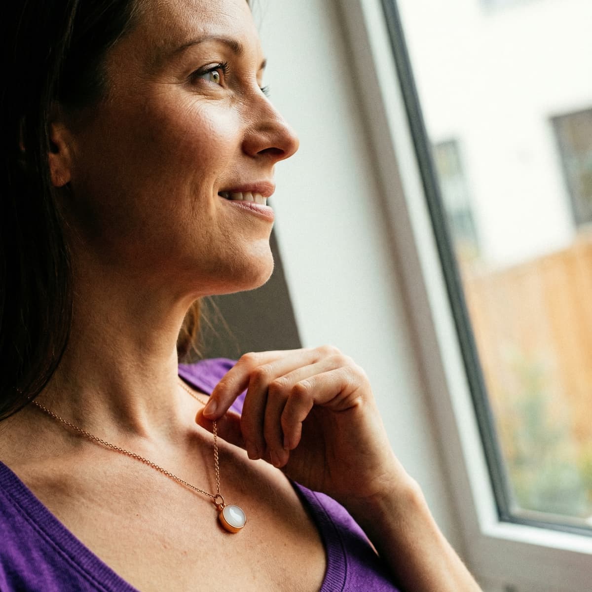 Woman standing by a window wearing a gold-toned keepsake pendant with a milky resin stone, made using a breastmilk necklace making kit.