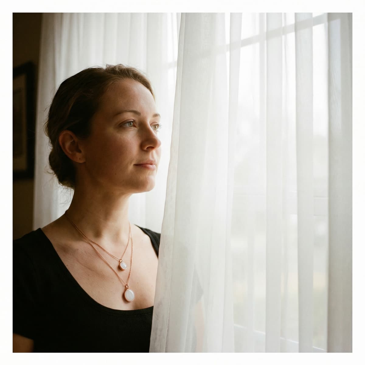 Woman wearing two breastmilk keepsake pendants and looking out a sunlit window, showcasing jewelry made with a breastmilk necklaces making kit.