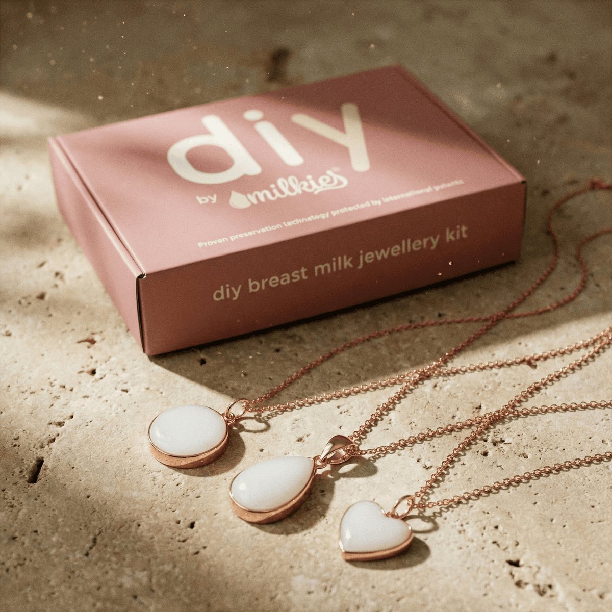 Rose-gold DIY by MILKIES pink box showing a breastmilk necklaces making kit with three white resin pendant necklaces (round, teardrop, and heart) arranged on a stone surface