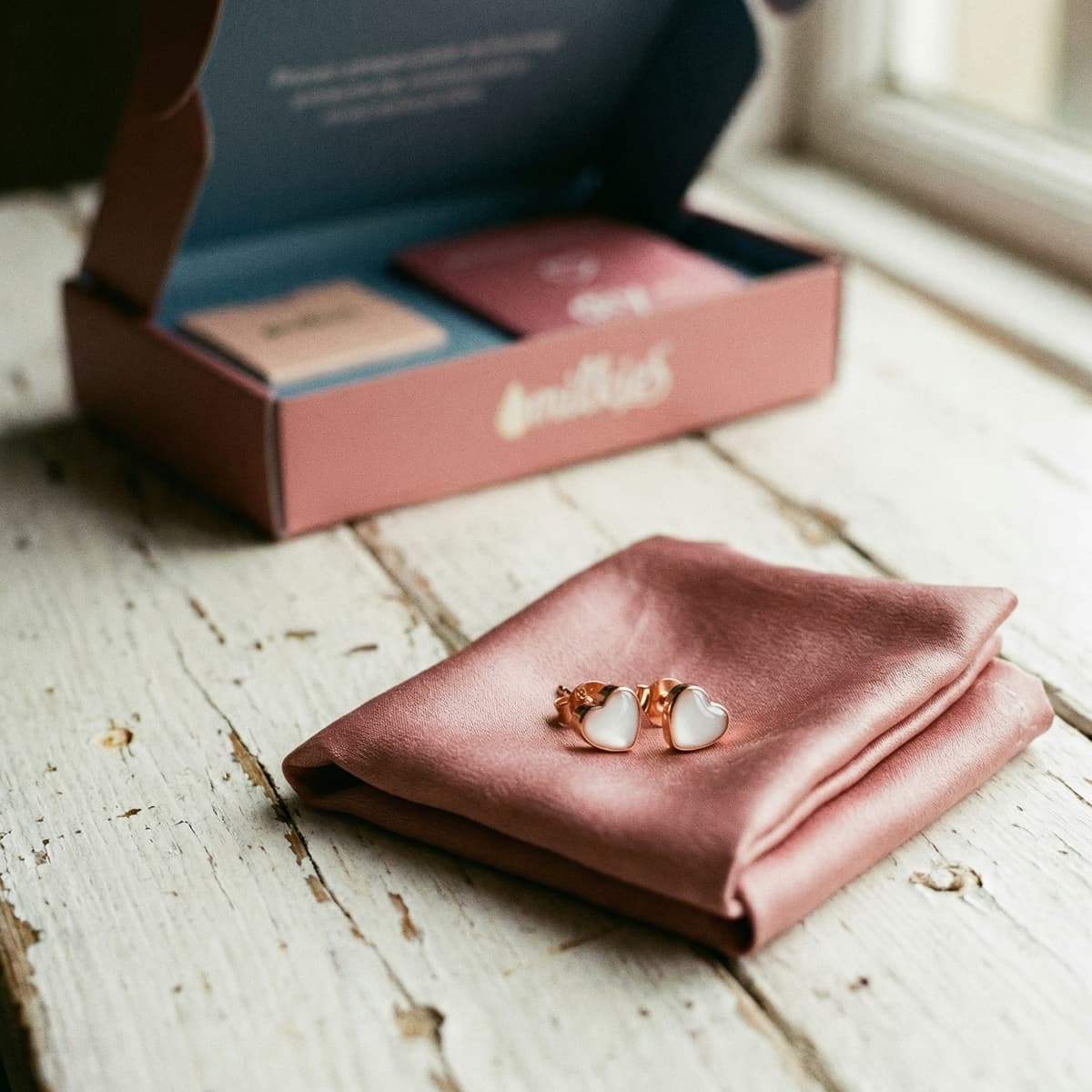 Heart-shaped rose gold stud earrings on a pink satin pouch next to an open DIY by MILKIES box, showing how a diy breastmilk earring making kit helps create personalized keepsake jewelry at home.