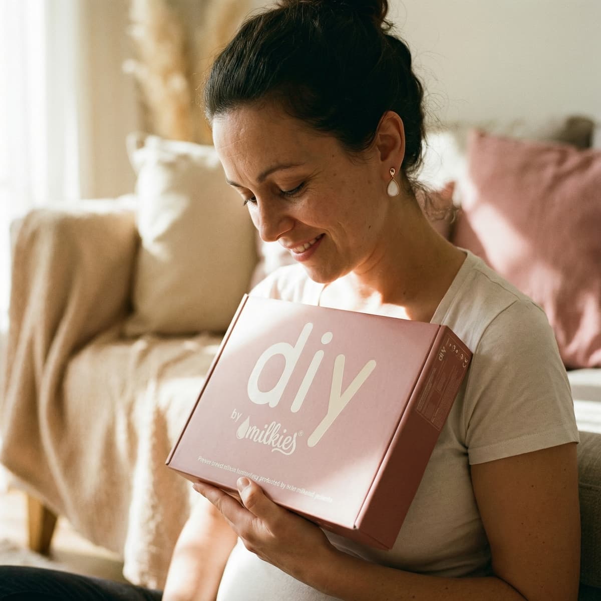 Smiling mom at home holding a pink DIY by MILKIES box, showing why a diy breastmilk earring making kit is a convenient, private way to create a meaningful keepsake.