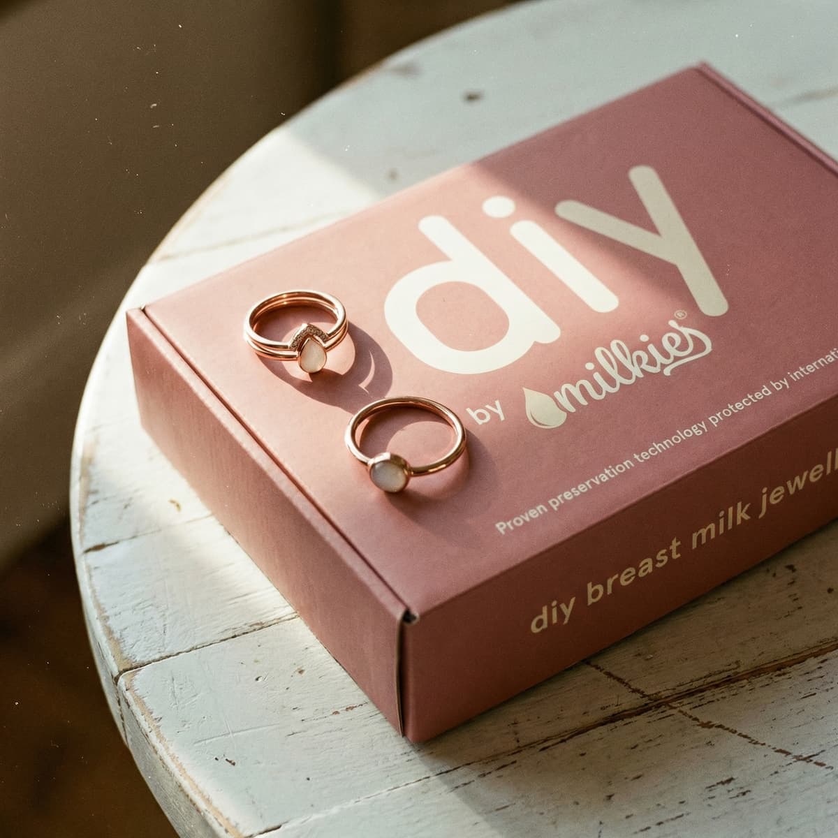 Rose-gold DIY by MILKIES jewelry kit showing diy breastmilk rings with milky white stones on a pink kit box, set on a rustic white table in warm natural light.