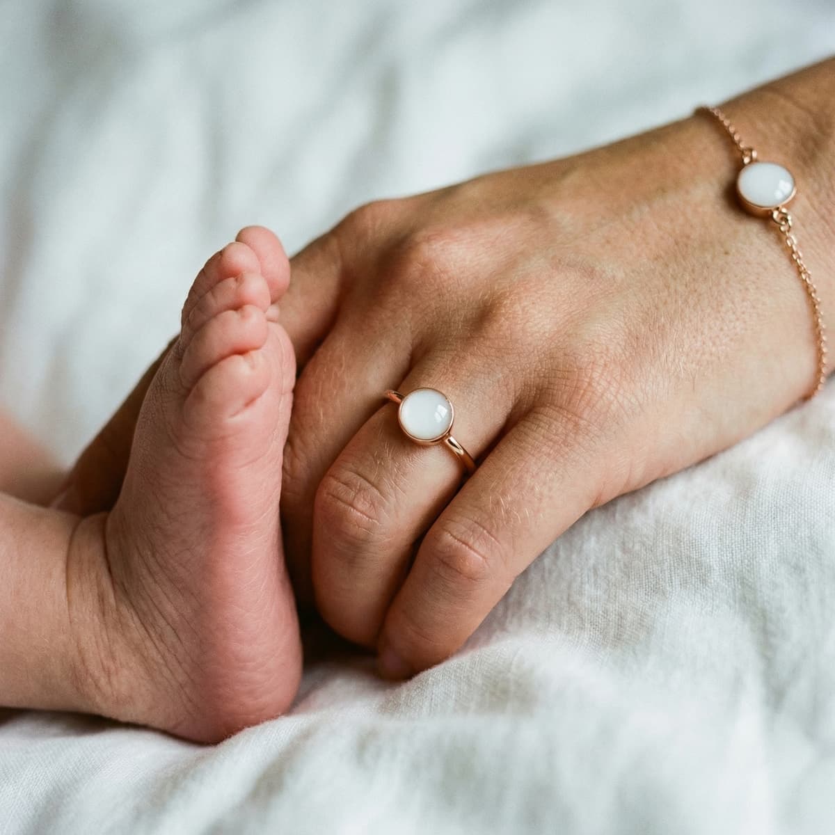 Mother holding baby’s tiny foot while wearing rose-gold ring and bracelet with milky white resin stones, showing how to make your own breastmilk jewelry at home.