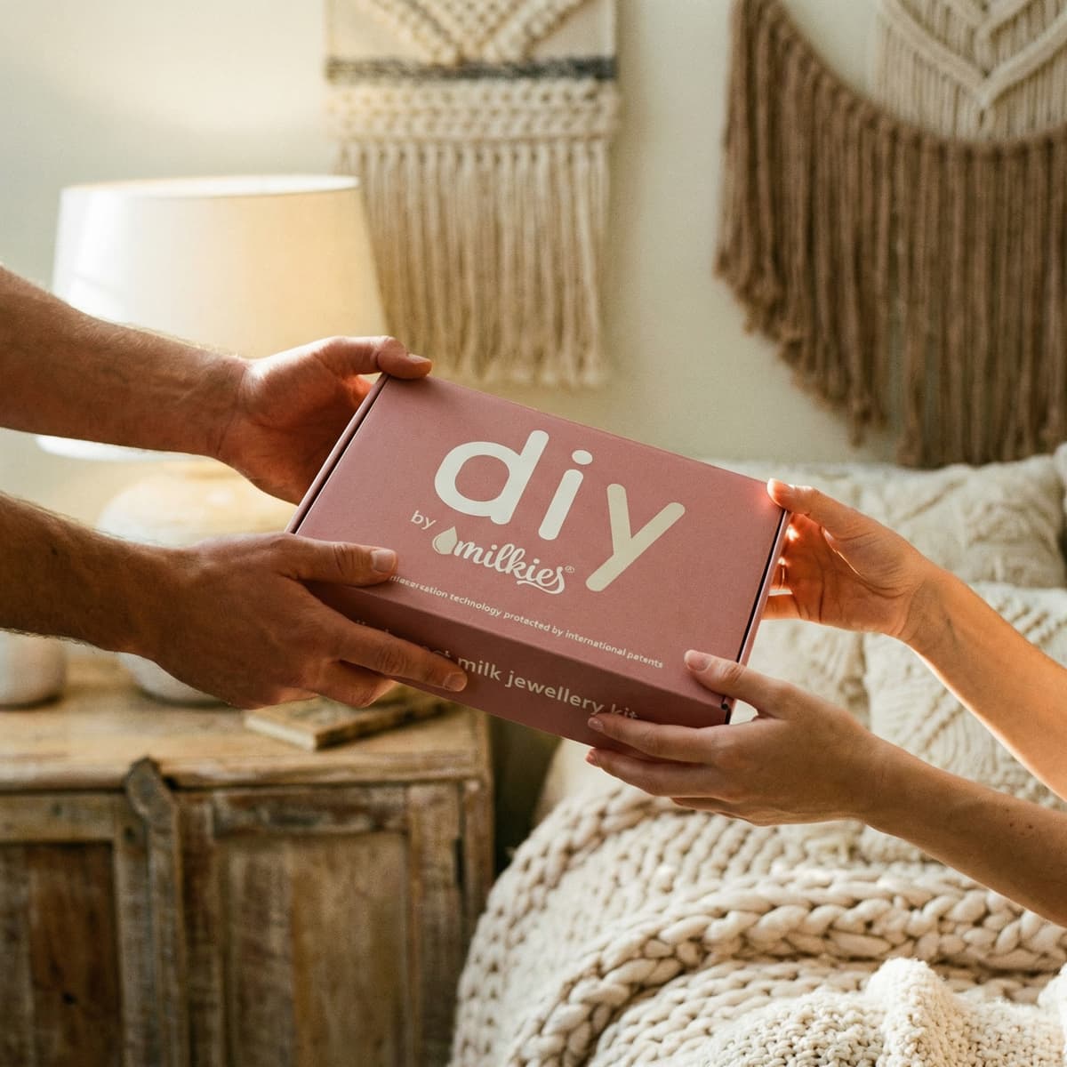 In a cozy bedroom, a partner hands over a pink DIY by MILKIES breastmilk jewellery kit box as a meaningful push present for a new mom.